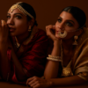 Two women adorned in traditional gold jewelry, wearing elegant sarees, showcasing ornate gold necklaces, bangles, earrings, and headpieces. The image promotes festive gold jewelry collections from Kanakpura, with a caption: "Celebrate with Gold Jewelry from Kanakpura."